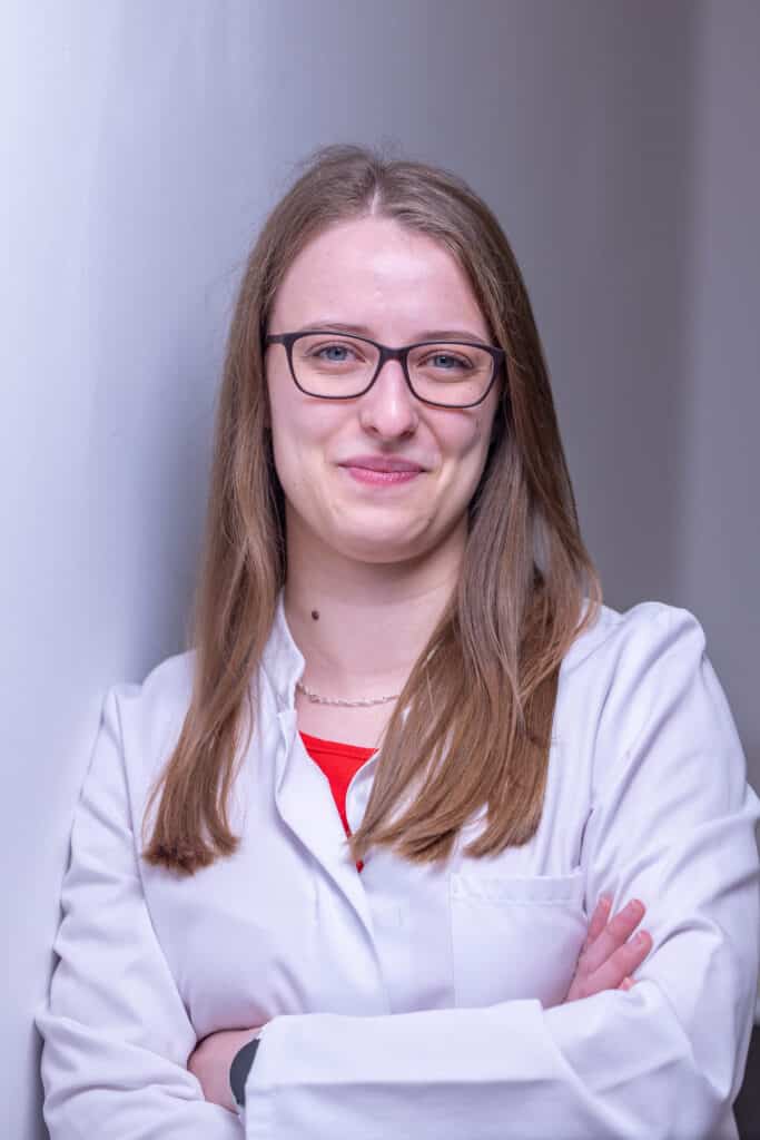 Frau mit langen Haaren und Brille, im weißen Laborkittel, steht mit verschränkten Armen und lächelt.