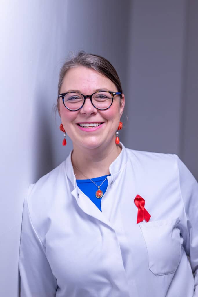 Frau in weißem Laborkittel mit roter Schleife, lächelnd, vor einer hellen Wand stehend.