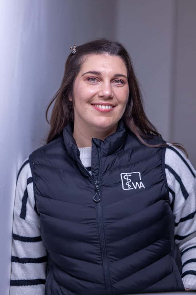 Eine Frau mit langen braunen Haaren, einem gestreiften Pullover und einer schwarzen Weste lehnt lächelnd an einer weißen Wand.